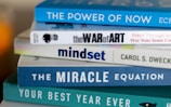 A stack of self-help and motivational books with colorful covers is neatly arranged. The titles include 'The Power of Now', 'The War of Art', 'Mindset', 'The Miracle Equation', and 'Your Best Year Ever'.