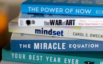 A stack of self-help and motivational books with colorful covers is neatly arranged. The titles include 'The Power of Now', 'The War of Art', 'Mindset', 'The Miracle Equation', and 'Your Best Year Ever'.