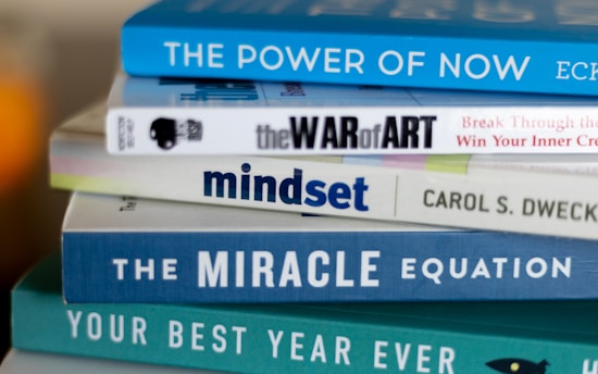 A stack of self-help and motivational books with colorful covers is neatly arranged. The titles include 'The Power of Now', 'The War of Art', 'Mindset', 'The Miracle Equation', and 'Your Best Year Ever'.