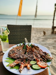A plate filled with grilled seafood including fish and prawns, garnished with lime, cucumber, and tomato slices, placed on a table near a beach setting. A refreshing drink with mint leaves, ice, and a lime slice is visible next to the plate. In the background, the sea and part of the sandy beach can be seen, along with a few beach flags.