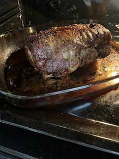 A large, well-roasted piece of meat sits in a glass baking dish inside an oven. The surface of the meat is caramelized and browned, indicating it is thoroughly cooked. The glass dish rests on an oven rack, and the oven interior is dark with a light illuminating the meat.