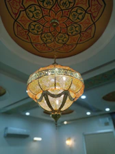 An intricately designed chandelier hangs from a ceiling with elaborate patterns. The chandelier is ornate, featuring a perforated design that allows light to shine through, casting patterns. The ceiling itself has a circular, mandala-like design in warm earthy tones.