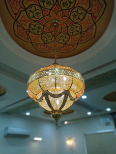 An intricately designed chandelier hangs from a ceiling with elaborate patterns. The chandelier is ornate, featuring a perforated design that allows light to shine through, casting patterns. The ceiling itself has a circular, mandala-like design in warm earthy tones.