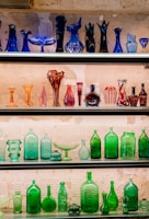 A display of various colorful glass bottles and vases arranged on shelves against a textured wall. The top shelf features dark blue and purple glassware in artistic shapes, while the middle shelf showcases red, pink, and orange vessels. The bottom shelves are filled with green glass bottles and dishes, creating a vibrant contrast.