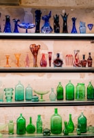 A display of various colorful glass bottles and vases arranged on shelves against a textured wall. The top shelf features dark blue and purple glassware in artistic shapes, while the middle shelf showcases red, pink, and orange vessels. The bottom shelves are filled with green glass bottles and dishes, creating a vibrant contrast.