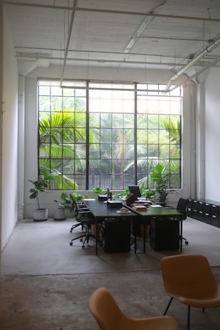 An industrial-style office space features large windows with grid patterns that allow ample natural light to illuminate the room. The view outside shows lush green foliage, contributing to a tranquil atmosphere. Inside, there are dark-colored desks arranged in a communal fashion with several office chairs, suggesting a collaborative workspace. Indoor plants in pots are placed strategically around the space, enhancing the natural aesthetic. The room's high ceilings and exposed pipes add an architectural element to the design.