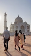 A group of people are walking towards the Taj Mahal, a stunning white marble mausoleum, as the sun sets in the background. The structure features its iconic dome and detailed architectural elements. The sky is clear with a soft glow.