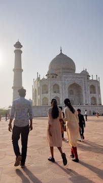 A group of people are walking towards the Taj Mahal, a stunning white marble mausoleum, as the sun sets in the background. The structure features its iconic dome and detailed architectural elements. The sky is clear with a soft glow.