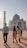 A group of people are walking towards the Taj Mahal, a stunning white marble mausoleum, as the sun sets in the background. The structure features its iconic dome and detailed architectural elements. The sky is clear with a soft glow.