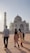 A group of people are walking towards the Taj Mahal, a stunning white marble mausoleum, as the sun sets in the background. The structure features its iconic dome and detailed architectural elements. The sky is clear with a soft glow.