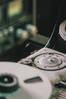 A close-up view of a vintage reel-to-reel tape recorder, featuring visible spools and knobs with metallic components. The image focuses on the mechanical details, capturing a sense of old-fashioned audio technology.