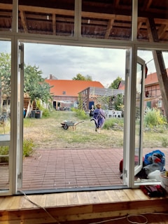 A view from indoors through large glass doors into a backyard setting. The foreground has a wooden floor leading to a patio with red tiles. Outside, there's grass, various plants, and shrubs. A person is running across the yard while pushing a wheelbarrow. In the background, there are buildings with red roofs and some chairs set up in the yard.