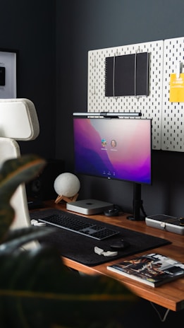 A modern home office setup featuring a sleek computer monitor with a purple gradient display, a compact computer unit, wireless keyboard and mouse on a dark felt mat, and a round desk lamp with a white spherical shade. The workspace includes a wooden desk and an upholstered chair. Above the desk, there are pegboards with a notebook and a yellow sticky note attached. A plant partially obscures the left side and there is a magazine on the desk.