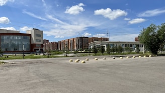 A spacious urban area with a large glass-fronted building on the left and a modern structure with a curved roof in the center. Residential buildings are visible in the background, and a line of bollards separates the paved area from the green space with trees and grass.