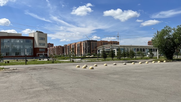 A spacious urban area with a large glass-fronted building on the left and a modern structure with a curved roof in the center. Residential buildings are visible in the background, and a line of bollards separates the paved area from the green space with trees and grass.