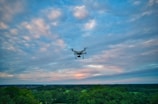 A drone flies over a lush, green forest with a backdrop of a sky filled with scattered clouds, displaying hues of blue and pink.