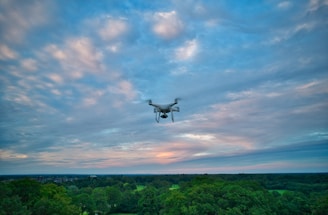 A drone flies over a lush, green forest with a backdrop of a sky filled with scattered clouds, displaying hues of blue and pink.