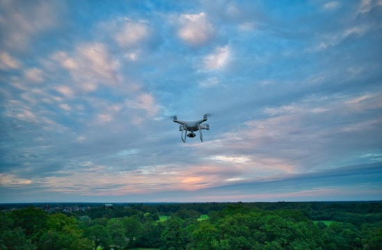 A drone flies over a lush, green forest with a backdrop of a sky filled with scattered clouds, displaying hues of blue and pink.