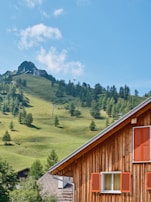 A rustic wooden house with orange shutters stands in the foreground, against a lush green hillside dotted with sparse trees. A mountainous peak rises in the background under a bright blue sky. Ski lift poles are visible along the slope.