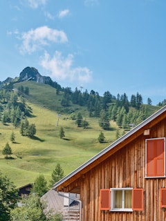A rustic wooden house with orange shutters stands in the foreground, against a lush green hillside dotted with sparse trees. A mountainous peak rises in the background under a bright blue sky. Ski lift poles are visible along the slope.