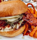 A close-up of a gourmet chicken sandwich featuring crispy fried chicken with a glossy red glaze. The sandwich includes pickles, lettuce, and a thick, toasted bun. Curled sweet potato fries accompany the sandwich on a plate.