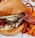 A close-up of a gourmet chicken sandwich featuring crispy fried chicken with a glossy red glaze. The sandwich includes pickles, lettuce, and a thick, toasted bun. Curled sweet potato fries accompany the sandwich on a plate.