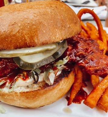 A close-up of a gourmet chicken sandwich featuring crispy fried chicken with a glossy red glaze. The sandwich includes pickles, lettuce, and a thick, toasted bun. Curled sweet potato fries accompany the sandwich on a plate.