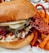 A close-up of a gourmet chicken sandwich featuring crispy fried chicken with a glossy red glaze. The sandwich includes pickles, lettuce, and a thick, toasted bun. Curled sweet potato fries accompany the sandwich on a plate.