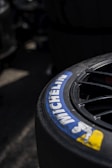 A close-up view of a Michelin tire with part of the brand logo and name clearly visible. The tire appears to be attached to a wheel with visible spokes, set against a dark and slightly blurred background.