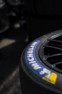 A close-up view of a Michelin tire with part of the brand logo and name clearly visible. The tire appears to be attached to a wheel with visible spokes, set against a dark and slightly blurred background.