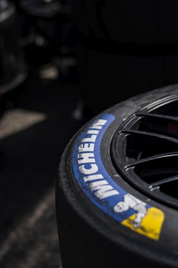 A close-up view of a Michelin tire with part of the brand logo and name clearly visible. The tire appears to be attached to a wheel with visible spokes, set against a dark and slightly blurred background.