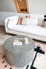 A modern living room featuring a white cushioned sofa adorned with patterned pillows against a light wall. In front of the couch, there is a round striped coffee table with a jar candle and a magazine on top. A plush, patterned rug covers the wooden floor, adding texture to the decor. A uniquely angled wooden shelf in the background contains a few small decorative items.