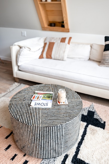 A modern living room featuring a white cushioned sofa adorned with patterned pillows against a light wall. In front of the couch, there is a round striped coffee table with a jar candle and a magazine on top. A plush, patterned rug covers the wooden floor, adding texture to the decor. A uniquely angled wooden shelf in the background contains a few small decorative items.