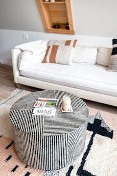 A modern living room featuring a white cushioned sofa adorned with patterned pillows against a light wall. In front of the couch, there is a round striped coffee table with a jar candle and a magazine on top. A plush, patterned rug covers the wooden floor, adding texture to the decor. A uniquely angled wooden shelf in the background contains a few small decorative items.