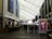 A covered walkway inside a large shopping or entertainment complex, with people walking along the path. On the sides are various storefronts and advertisements. Banners and digital screens display promotional content.