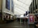 A covered walkway inside a large shopping or entertainment complex, with people walking along the path. On the sides are various storefronts and advertisements. Banners and digital screens display promotional content.