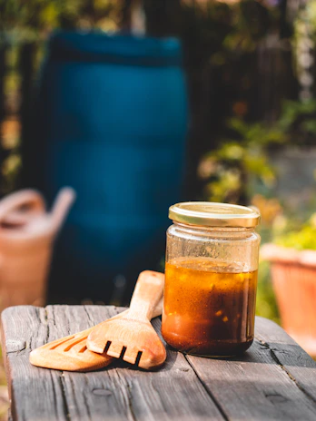 A glass jar with a golden lid containing a brown liquid is placed on a wooden surface. Next to it, there are wooden kitchen utensils. The background features a blurred garden scene with greenery and a blue water barrel.