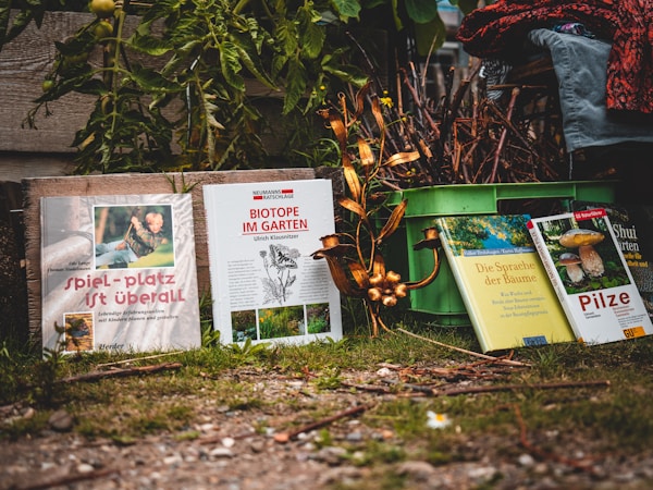 A collection of books is arranged in a garden setting, with lush greenery in the background. The books have colorful covers and are resting on a grassy patch, suggesting themes related to nature and gardening.