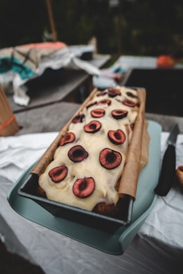 A loaf cake topped with a cream-like frosting and decorated with halved cherries is presented in a rectangular baking tray. The background includes a table covered with a white cloth and some other indistinct objects, giving an informal, cozy setting.
