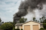 A house with a beige exterior and two garage doors is surrounded by green trees and bushes. Dark black smoke billows dramatically into the sky, suggesting a fire nearby.