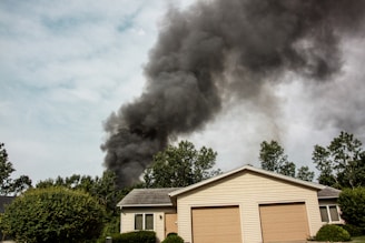 A house with a beige exterior and two garage doors is surrounded by green trees and bushes. Dark black smoke billows dramatically into the sky, suggesting a fire nearby.