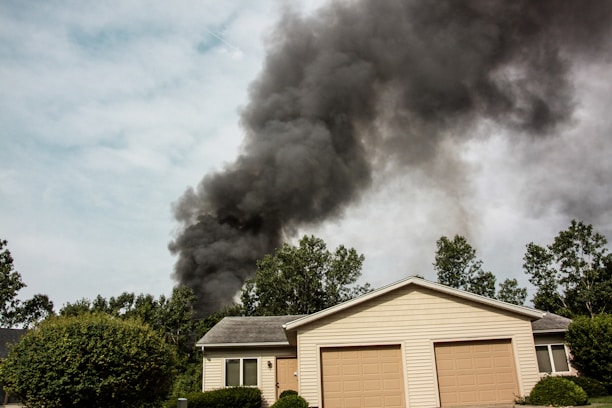 A house with a beige exterior and two garage doors is surrounded by green trees and bushes. Dark black smoke billows dramatically into the sky, suggesting a fire nearby.
