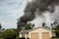 A house with a beige exterior and two garage doors is surrounded by green trees and bushes. Dark black smoke billows dramatically into the sky, suggesting a fire nearby.