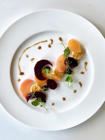 A minimalist presentation of a gourmet dish on a white plate. The arrangement includes slices of beets and other root vegetables in varying shades of pink and orange. Garnished with small leaves and pea shoots, with a drizzle of sauce artfully placed around the vegetables.