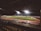 A nighttime football match is being played in a stadium illuminated by bright floodlights. The field is green, and a track surrounds it, with seating areas visible. Spectators can be seen watching the game from the stands.
