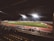 A nighttime football match is being played in a stadium illuminated by bright floodlights. The field is green, and a track surrounds it, with seating areas visible. Spectators can be seen watching the game from the stands.