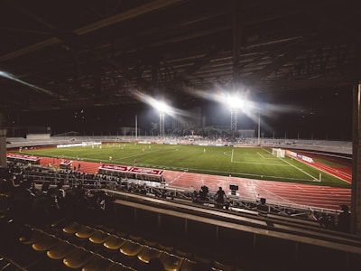 A nighttime football match is being played in a stadium illuminated by bright floodlights. The field is green, and a track surrounds it, with seating areas visible. Spectators can be seen watching the game from the stands.