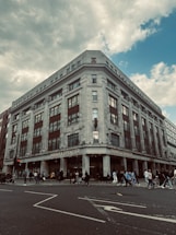 A large, multi-story retail building with a classic architectural style, featuring tall windows and decorative elements. The store's name, Marks & Spencer, is prominently displayed at the top of the building. In front of the building, numerous people are walking along the sidewalk, suggesting a busy urban environment. The sky above is partly cloudy.
