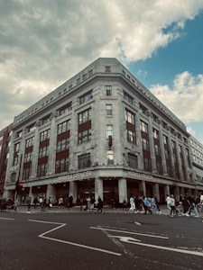 A large, multi-story retail building with a classic architectural style, featuring tall windows and decorative elements. The store's name, Marks & Spencer, is prominently displayed at the top of the building. In front of the building, numerous people are walking along the sidewalk, suggesting a busy urban environment. The sky above is partly cloudy.