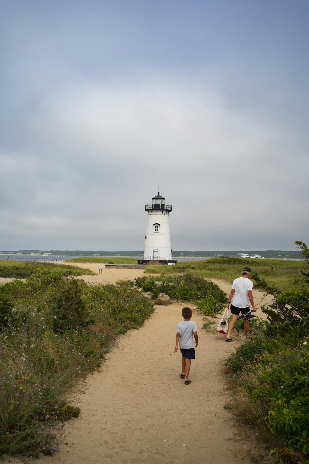 a couple of people walking towards a lighthouse on a path by a body of water